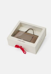 Accessorio Calvin Klein in una presentazione in scatola. Include una borsa beige con un accento a motivo, chiusura con zip e un nastro rosso.