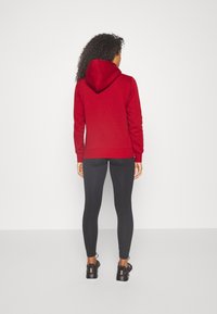 Červená mikina s kapucí a klokaní kapsou, spárovaná s černými legínami a černými sportovními botami. Tkanina vypadá jemně a strukturovaně.