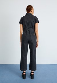 Svart denim jumpsuit med korte ermer, innsnevret midje og vide bukser. Har to baklommer og et minimalistisk design.