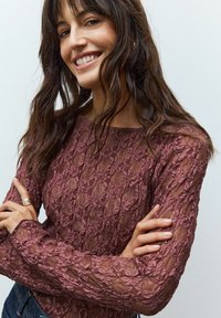 Lace top met lange mouwen in mauve, featuring een bloemenpatroon met een iets doorschijnende textuur en een ronde halslijn, gecombineerd met blauwe jeans.