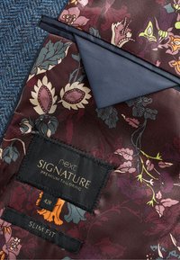 Étiquette Next Signature Premium Tailoring sur un tissu bordeaux foncé avec des motifs floraux dans des nuances de rose, vert et jaune. Coupe ajustée, taille 42R.
