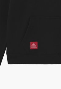 Haut à capuche noir en fabric doux avec une poche avant et des poignets côtelés. Présente une étiquette rouge avec un texte blanc en bas à droite.