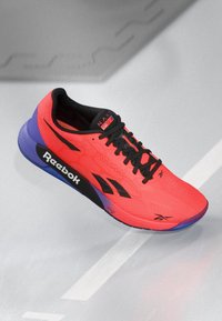 Pantofii de alergare roșii strălucitori cu un material texturat, accente negre și talpă violet. Prezintă logo-ul Reebok și șireturi negre. Designul este ușor.