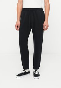 Černé joggingové kalhoty s elastickým pasem, nastavitelným šněrováním a zúženými kotníky. Stylizované s černými teniskami a bílými ponožkami.