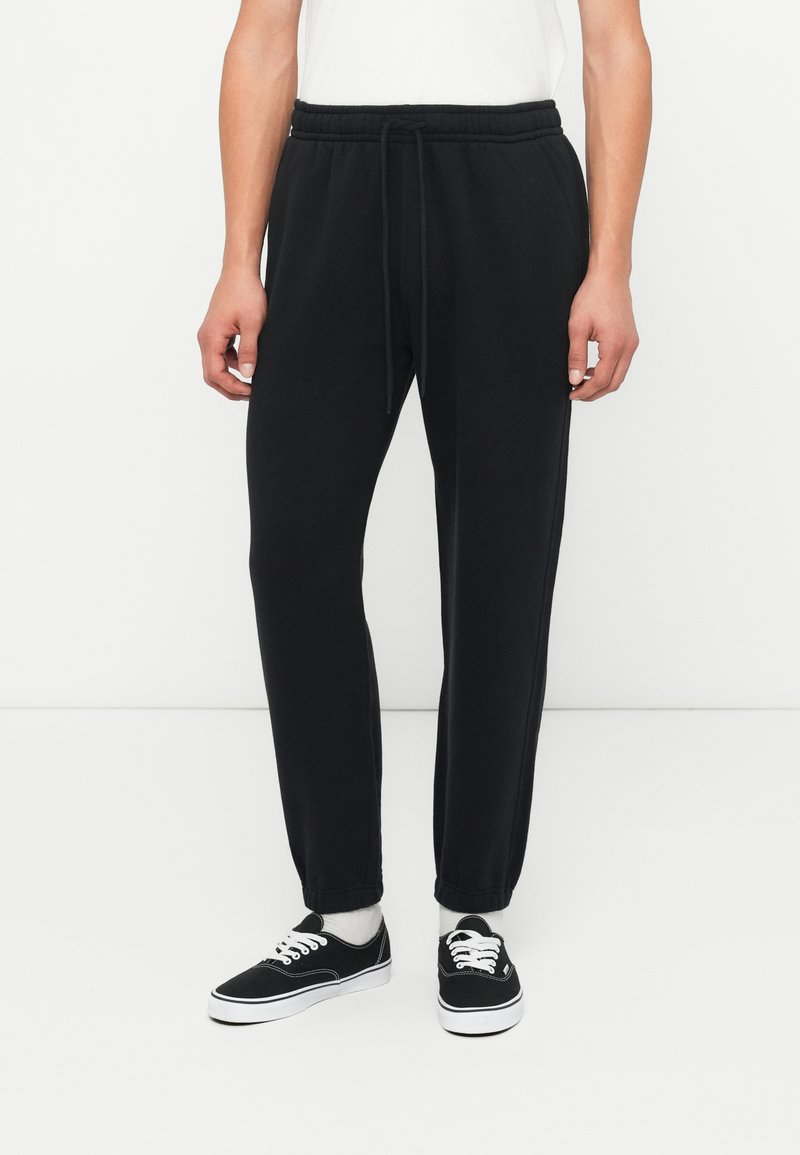 Černé joggingové kalhoty s elastickým pasem, nastavitelným šněrováním a zúženými kotníky. Stylizované s černými teniskami a bílými ponožkami.
