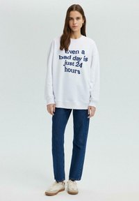 Witte sweatshirt met marineblauwe tekst "Zelfs een slechte dag is slechts 24 uur," gecombineerd met blauwe jeans en neutrale sneakers, staand tegen eenlichte achtergrond.