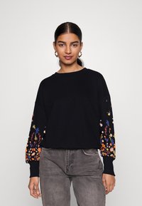 Μαύρη ζακέτα με κεντημένα λουλουδάτα σχέδια σε ζωντανά χρώματα στα μανίκια. Χαλαρή εφαρμογή με στρογγυλή λαιμόκοψη και ραμμένα μανίκια.