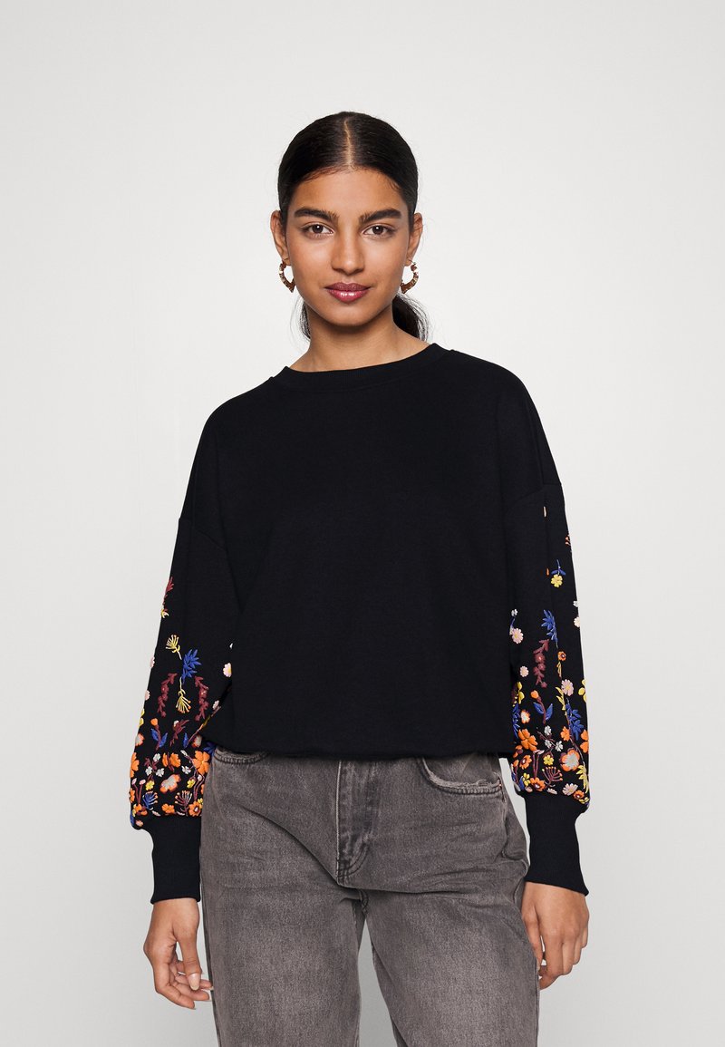 Μαύρη ζακέτα με κεντημένα λουλουδάτα σχέδια σε ζωντανά χρώματα στα μανίκια. Χαλαρή εφαρμογή με στρογγυλή λαιμόκοψη και ραμμένα μανίκια.