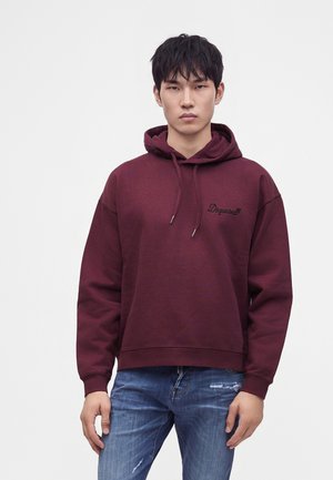 Jeune homme aux cheveux courts noirs portant un sweat à capuche bordeaux et un jean bleu déchiré, debout devant un fond clair uni.