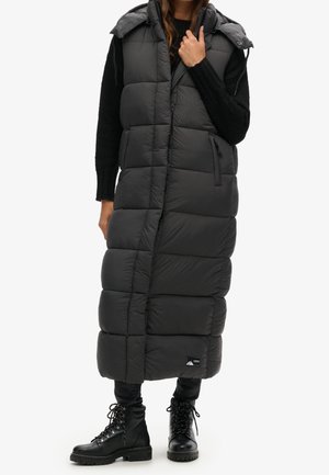 Femme portant un long manteau sans manches noir matelassé avec capuche, pull noir, pantalon noir et bottes noires à lacets, debout sur fond blanc.