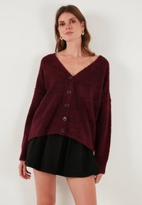 Burgundy színű bolyhos kardigán V-nyakkal, hosszú ujjakal és hat elülső gombbal. Az alja elöl rövidebb, hátul hosszabb. Texturált anyag.