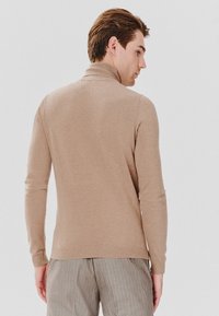 Beżowy sweter z golfem o dopasowanym kroju, z ściągaczami na rękawach i u dołu. Materiał wydaje się miękki i teksturowany, noszony z szaro-paskowanymi spodniami.