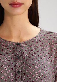 Blouse grise avec un motif floral en rose et violet, col rond et cinq boutons noirs sur le devant. Tissu texturé.