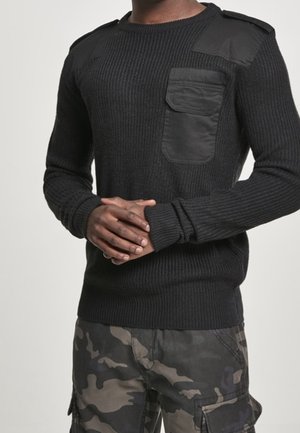 Mężczyzna w czarnym, dzianinowym swetrze z łatami na ramionach i klatce piersiowej oraz w szaro-czarnych spodniach typu cargo w kamuflażu, stojący ze złączonymi dłońmi.