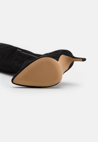 Bota de tacón alto de ante negro con punta afilada, un detalle de cremallera suave en el lateral y una suela de goma texturizada en color beige.