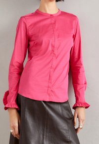 Mujer vistiendo una blusa de manga larga rosa brillante con puños acampanados y una falda de cuero negra, con las manos descansando a los lados.