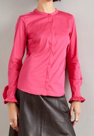 Vrouw in een felroze blouse met lange mouwen en gerimpelde manchetten, gecombineerd met een zwarte leren rok, met haar handen langs haar lichaam.