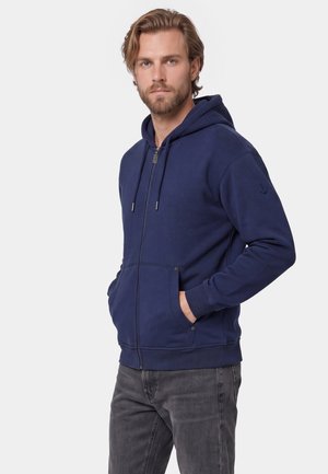 Mand med skæg iført en mørkeblå hættetrøje med lynlås med hænderne i frontlommerne og grå jeans mod en ensartet lys baggrund.