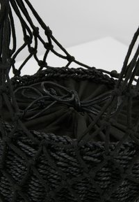 Panier en macramé noir tissé avec un doublure en tissu à cordon à l'intérieur, sur une surface blanche contre un fond blanc.