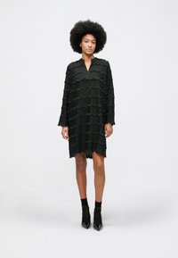 Mulher com um penteado afro natural, vestindo um vestido longo preto texturizado de mangas compridas e meias ankle pretas com sapatos de salto agulha.