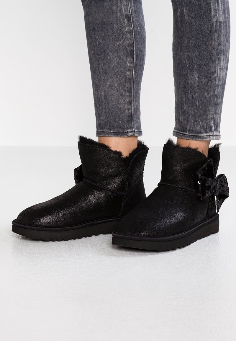 Bottines noires à la cheville avec doublure duveteuse et détail nœud en sequins, portées avec un jean skinny gris délavé.