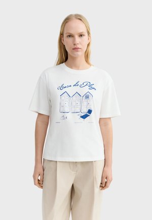Kobieta nosząca białą koszulkę z niebieskimi ilustracjami trzech domków plażowych, leżaka i napisem „Casa de Playa.”