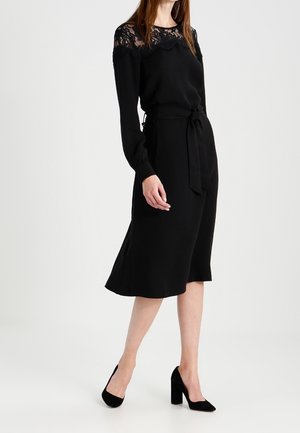 Zwarte jurk met een kanten halslijn, lange mouwen en een gestrikte taille. De rok is aangesloten bij de taille en loopt uit tot halverwege de kuit.