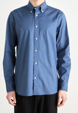 Mann trägt ein marineblaues Hemd mit langen Ärmeln und Knöpfen und kleinem Logo auf der Brust, kombiniert mit schwarzer Hose, steht vor schlichtem Hintergrund.