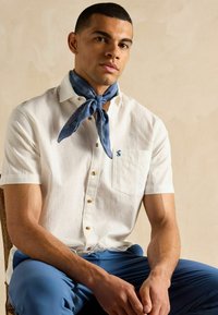 Balta īsām piedurknēm krekli ar brūniem pogām un krūšu kabatu, apvienoti ar zilām biksēm un zilu rakstainu bandanu ap kaklu.