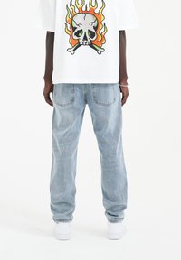 T-shirt grafica bianca con design di teschio e fiamme, abbinata a jeans in denim azzurro chiaro, caratterizzati da una vestibilità rilassata e orli risvoltati.