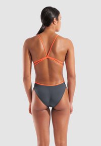 Vrouw met donker haar in een lage paardenstaart, draagt een grijze hoog uitgesneden bikini broek en een oranje bikini top met bandjes, gezien vanaf de achterkant.