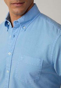 Light blue button-up shirt with a collar, single chest pocket, and subtle texture. Features small white buttons and an embroidered detail on pocket.