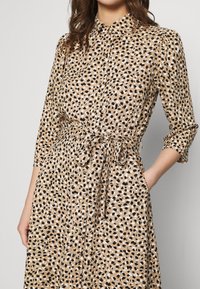 Femme portant une robe boutonnée tachetée de marron, noir et blanc avec une ceinture nouée à la taille et des manches trois-quarts, la main dans la poche.