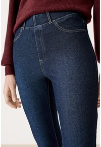 Tmavě modré džínové kalhoty s hladkou strukturou, vysokým pasem a výrazným žlutým prošíváním. Oděv je doplněn vínovým svetrem.