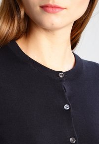 Mörkblå cardigan med rund halsringning, prydd med en rad knappar i kontrasterande färg och en slät stickad textur.