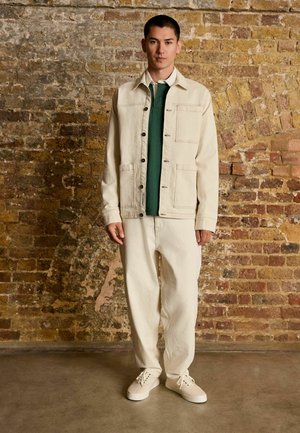 Beige denim jacket over a green shirt, paired with beige wide-leg pants and white sneakers. Brick wall background.