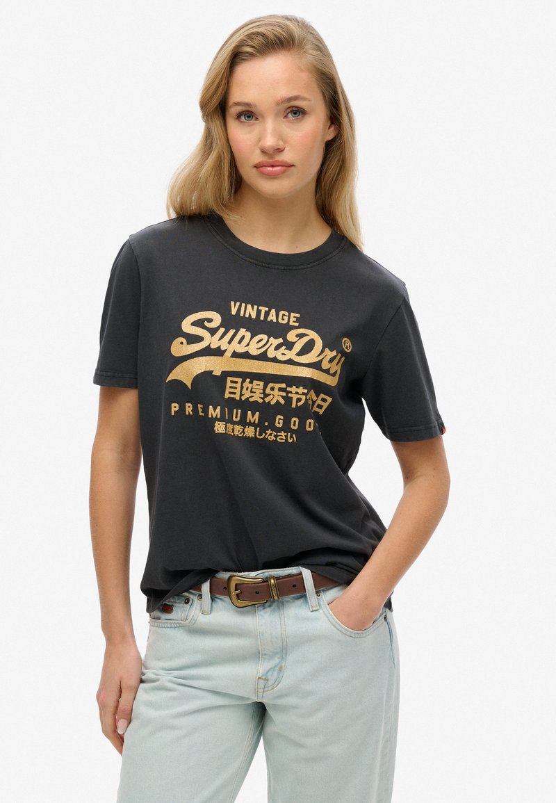 Zwarte kortemouwen T-shirt van katoen, met gouden vintage-stijl grafische tekst; gedragen met lichtblauwe jeans en een brown riem.