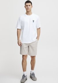 Hvit t-skjorte med kort erm og en liten brodert design, sammen med beige shorts og grå joggesko. Enkel, uformell antrekk.