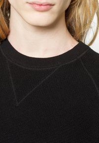 Gros plan sur la partie inférieure du visage et le cou d’une personne portant un pull noir texturé à col rond, avec une peau claire et des cheveux blond fraise.