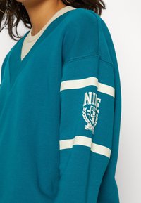 Teal tröja med V-ringning, kontrasterande krämfärgade detaljer på ärmarna och en tryckt logo med lagerblad detaljer på högra ärmen.