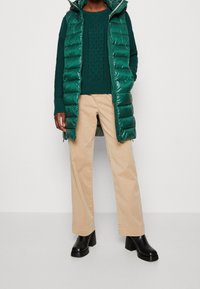 Veste matelassée verte avec capuche portée sur un pull vert foncé texturé, associée à un pantalon beige à jambes larges et des bottines noires à talons épais.