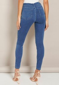Feszes kék farmernadrág elasztikus denim anyagból, magas derékkal, két hátsó zsebbel és sima textúrával. Meztelen színű, pántos magassarkúval párosítva.