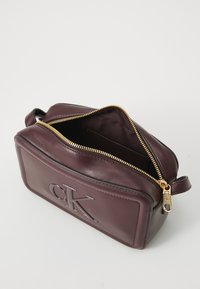 Pochette en cuir bordeaux avec une fermeture éclair dorée, logo "CK" gaufré sur le devant et une texture lisse. L'intérieur est doublé en tissu.