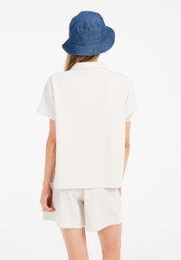 Weißes kurzärmliges Hemd mit strukturiertem Muster, kombiniert mit weißen Shorts. Ein blauer Denim-Eimerhut sorgt für Farbe und Kontrast.