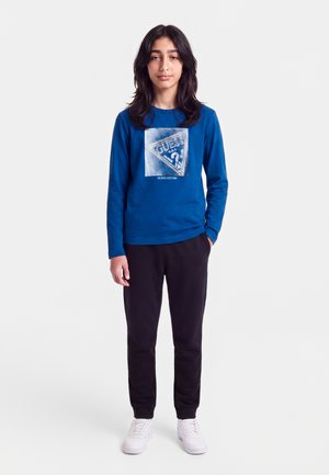 Jeune personne aux longs cheveux foncés portant un t-shirt Guess bleu à manches longues, un pantalon noir et des baskets blanches, debout avec une main dans la poche.