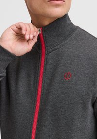 Hall hall zipiga jope kõrge kraega. Omab punast lukuosa ja väikest punast logo. Materjal tundub pehme ja sileda tekstuuriga.