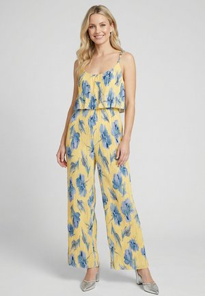 Femme souriante portant une combinaison sans manches jaune avec un imprimé floral bleu et des chaussures à talons argentés, debout devant un fond uni.