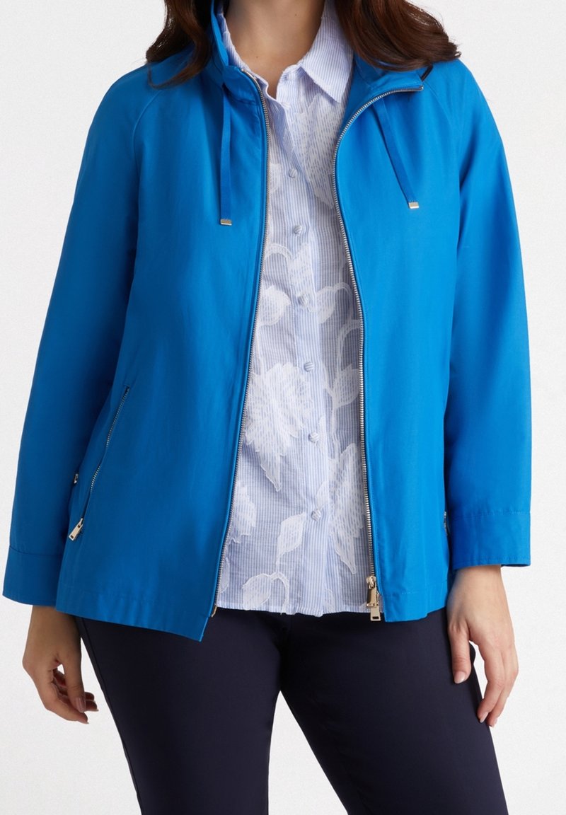Niebieska lekka kurtka z kapturem, posiadająca zamek błyskawiczny na całej długości z przodu, dwie zasuwane boczne kieszenie oraz gładką fakturę. Nałożona na białą bluzkę w kwiaty.