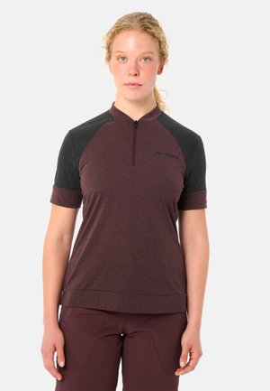 Junge Frau mit zusammengebundenem blondem Haar, die ein kurzärmeliges, bordeauxrotes und dunkelgraues Sportshirt mit Reißverschluss sowie passende Hose trägt.