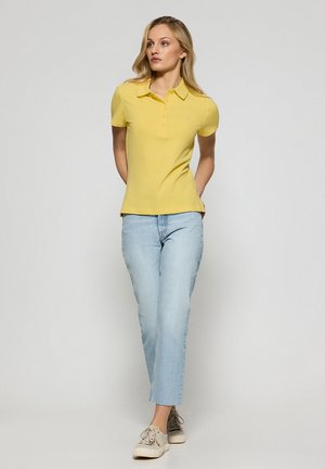 Polo shirt giallo, maniche corte, design aderente, jeans azzurri chiari e sneakers beige. Colletto semplice e dettaglio con bottoni sulla maglietta.